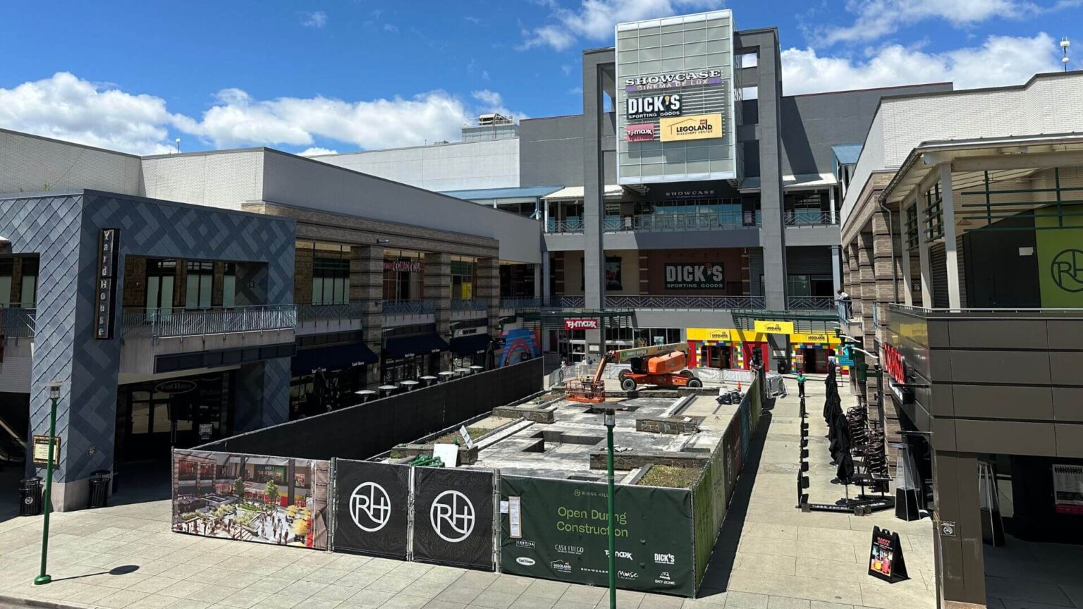 A shopping mall with multiple storefronts, including a construction site in the center surrounded by safety barriers and a "COMING SOON" banner.